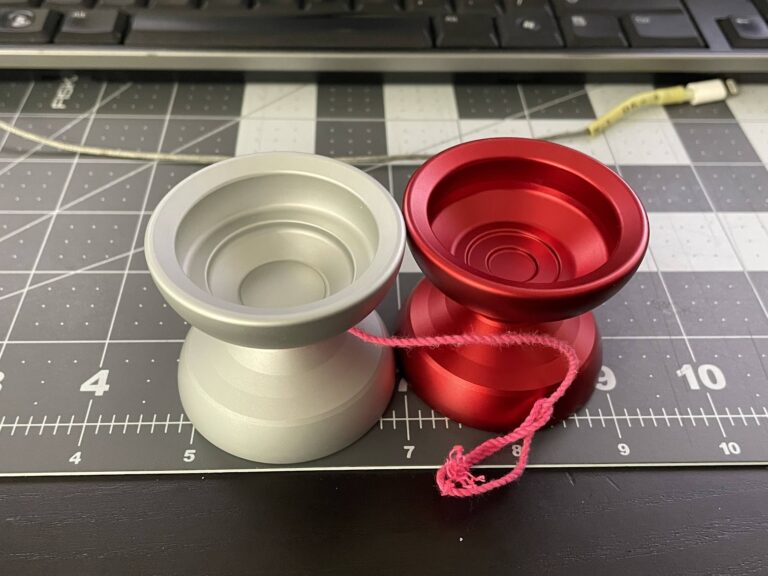 A silver yoyo prototype next to a red  yoyo prototype on a cutting mat, in front of a keyboard and USB cable.